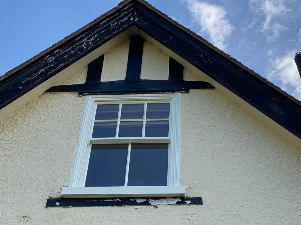 Close-up of an upper house window with peeling paint on the trim and textured beige exterior walls under a gabled roof—an ideal spot to repair fascia and soffit for improved curb appeal.