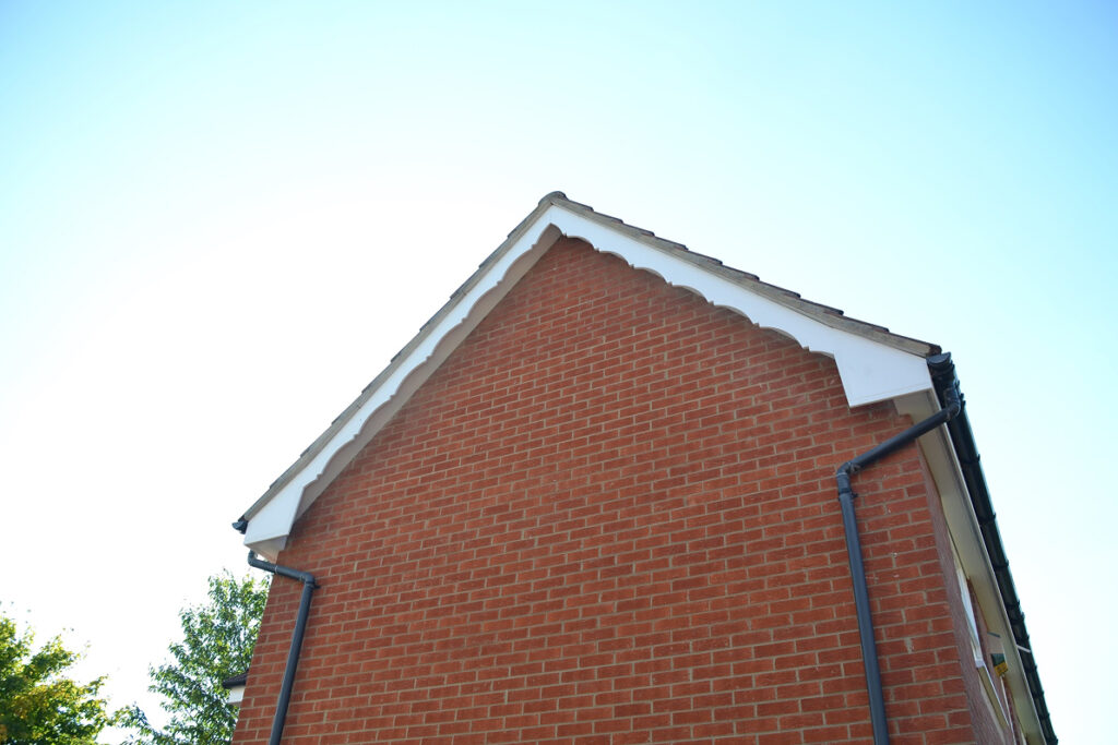 The gable end of a red brick house with scalloped white trim and black rain gutters under a clear blue sky, showcasing recently repair fascia and soffit for a polished finish.