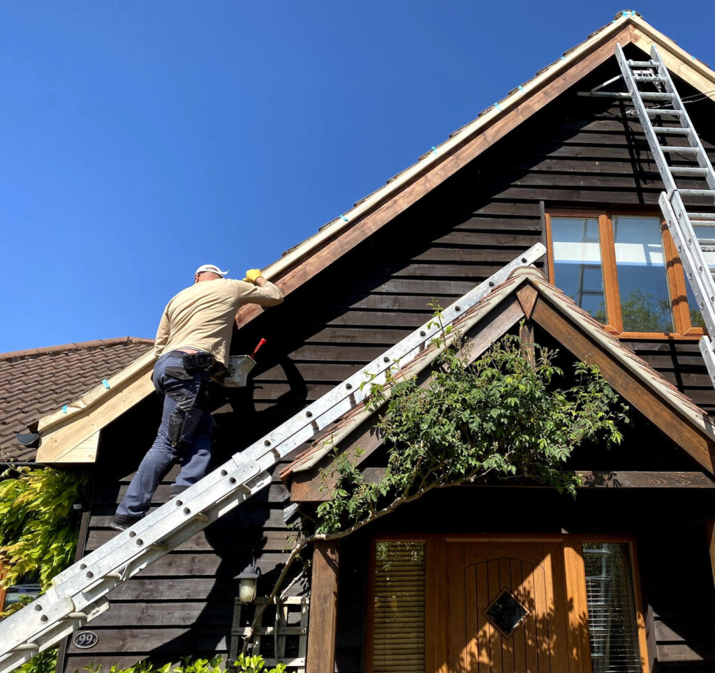 A person stands on a ladder working on the roof trim of a dark wooden house under clear blue skies.