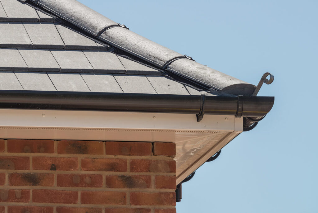 Close-up of a house roof corner with black slates, a black metal gutter, and brick wall against a clear blue sky—ideal for those looking to repair fascia and soffit.