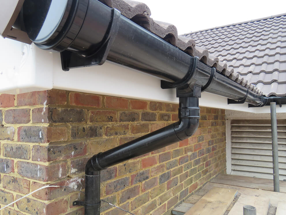 Black plastic guttering and downpipe system fitted to the edge of a tiled roof on a brick house exterior, ideal when you need to repair fascia and soffit for long-lasting protection.
