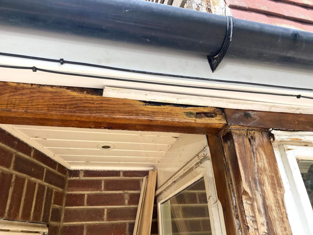 Close-up of an exterior wooden beam under a roof showing signs of water damage and rot, with some boards removed and insulation exposed.