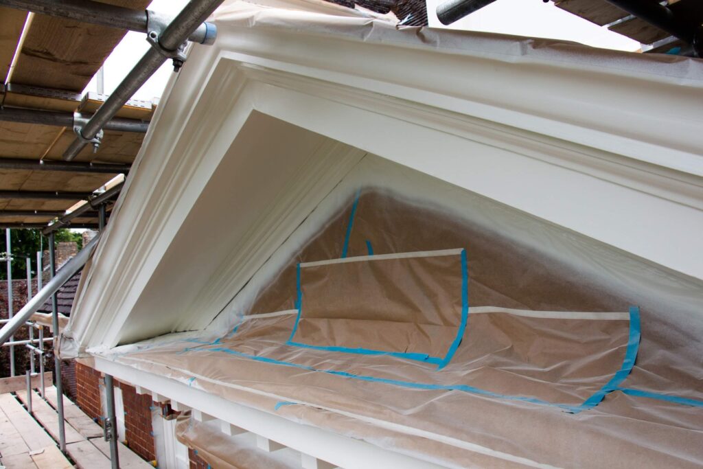 Painted architectural trim and a dormer window covered with masking paper and tape, surrounded by scaffolding for construction or renovation work.