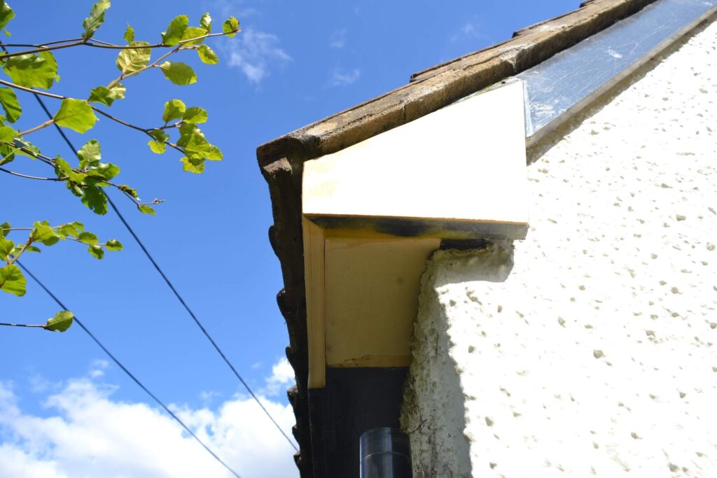 Close-up view of the corner of a house roof with textured white walls, blue sky, tree branches, and utility wires in the background.