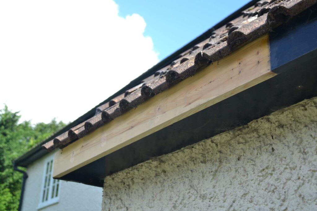 Close-up of a house roof edge showing new wooden fascia board installed beneath brown roof tiles above a textured exterior wall.