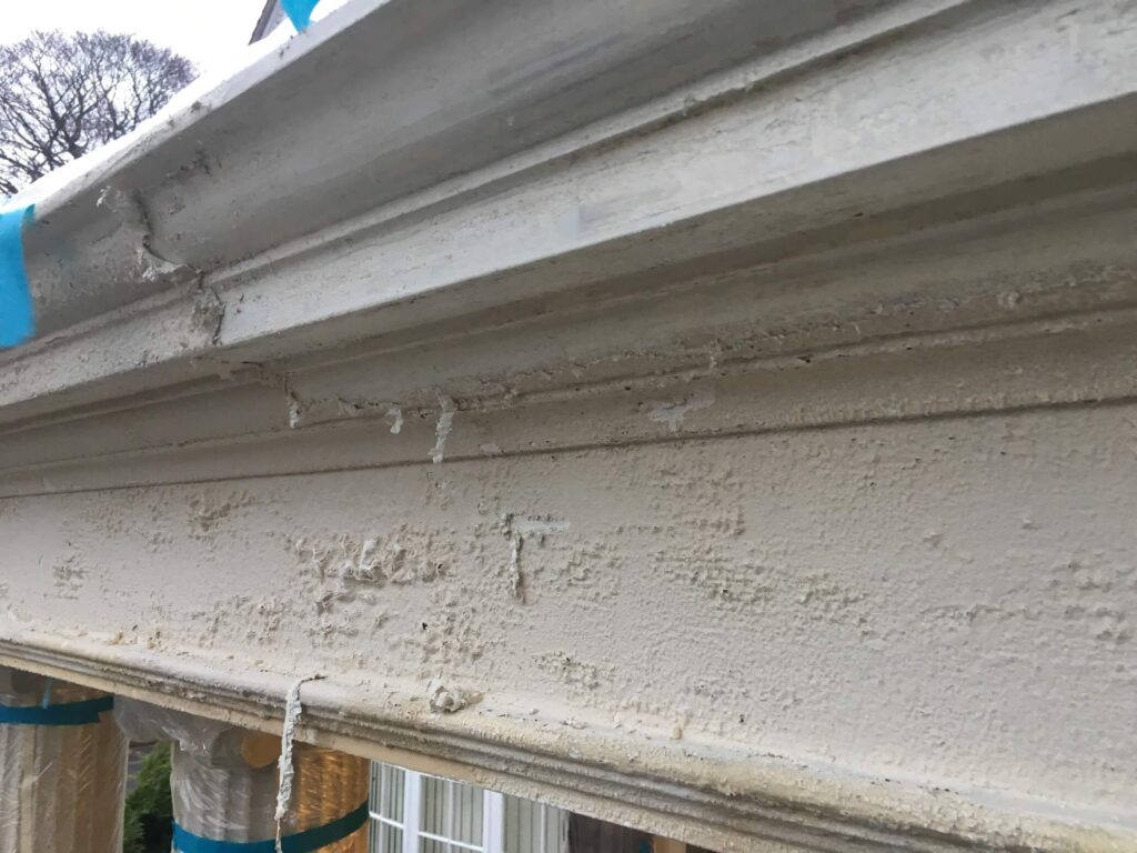 Close-up of a weathered and peeling exterior cornice, showing cracked and flaking white paint with some areas revealing damage to the underlying surface.