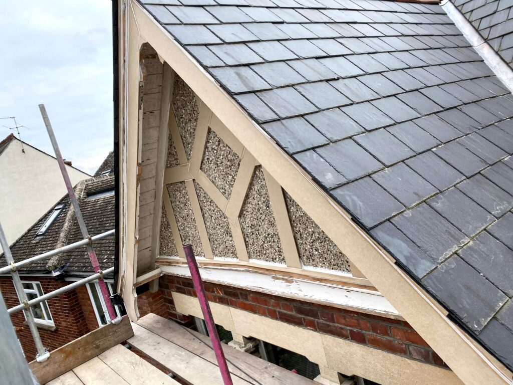 A close-up view of a house roof under renovation, showing slate tiles, exposed timber framing, pebble dash infill, scaffolding, and work underway to repair fascia and soffit.