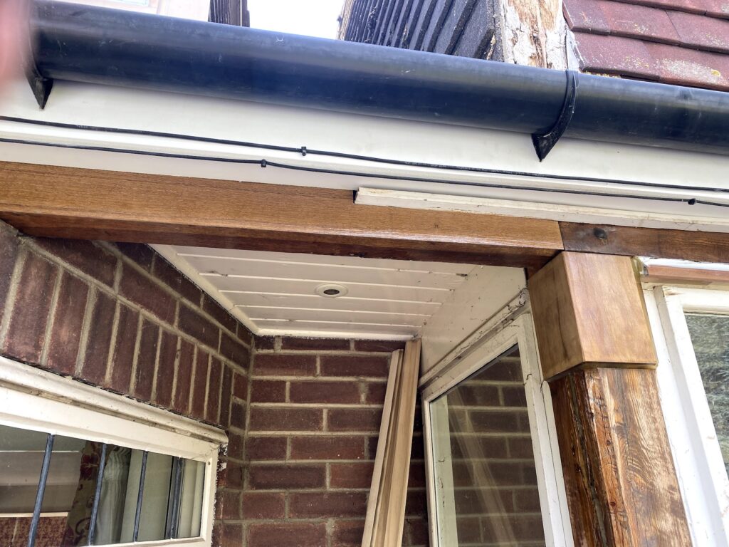 View of a brick patio corner with a white soffit ceiling, wooden beams, a black gutter, and windows with white frames. Several wooden planks lean against the wall.