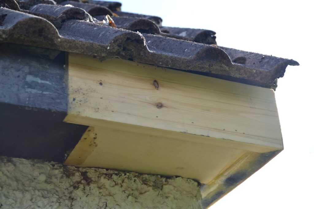 A close-up view of a house roof corner with a new wooden fascia board installed beneath old, weathered roof tiles.