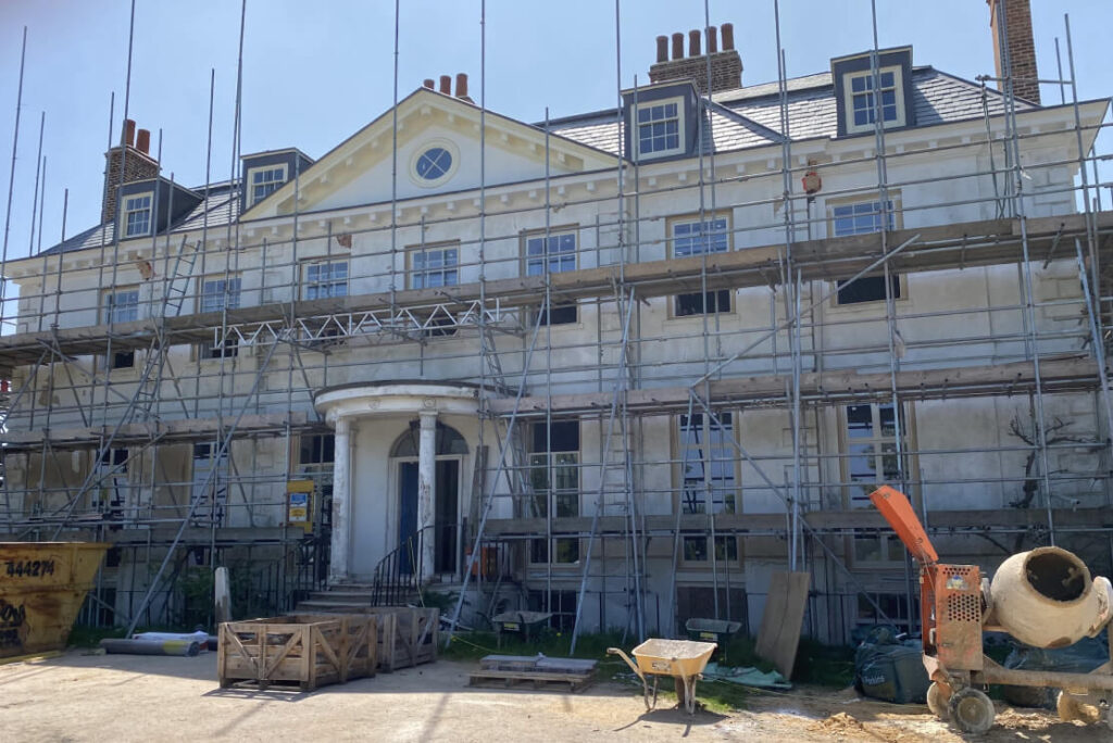 Large house under renovation, with scaffolding and construction equipment all around; workers on-site to repair fascia and soffit, while a skip and cement mixer sit out front.